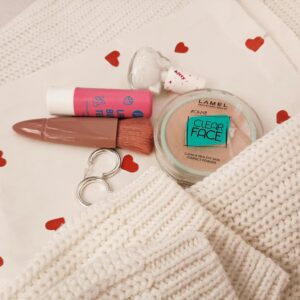 Stylish flat lay featuring makeup items, white knit, and heart-patterned cloth for beauty inspiration.