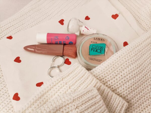 Stylish flat lay featuring makeup items, white knit, and heart-patterned cloth for beauty inspiration.
