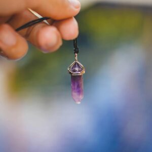 Home Close-up of a hand holding a purple amethyst pendant against a blurred outdoor background.